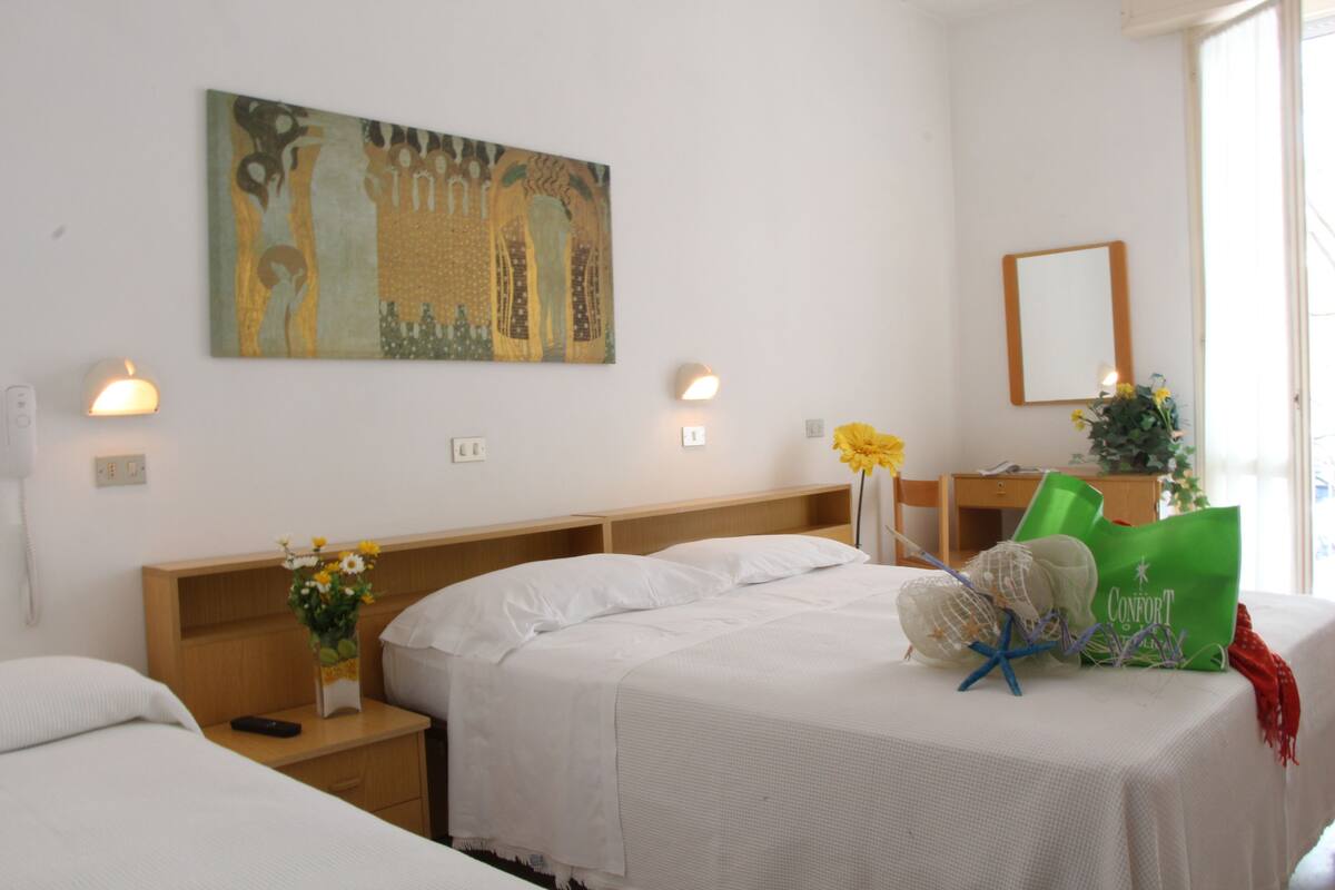 A hotel room with two beds, a painting on the wall, bedside lamps, a vase with flowers, and a green bag on the table.