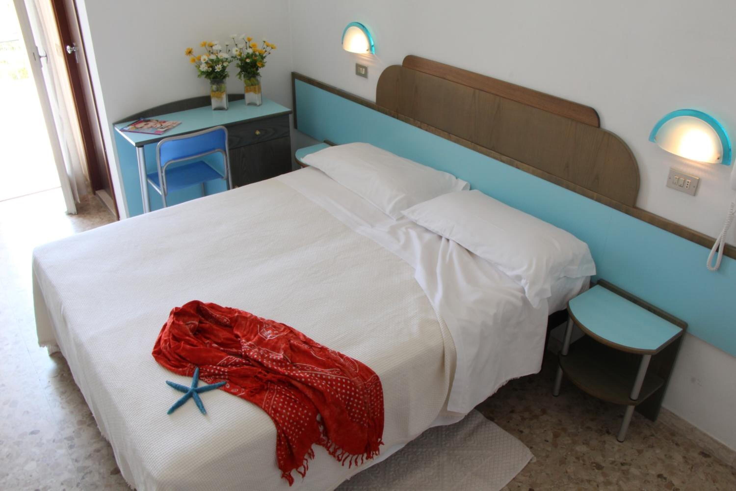 Una cama con sábanas blancas, una manta roja y una decoración de estrella de mar azul. Hay dos mesitas de noche con lámparas, un escritorio con una silla y un jarrón con flores en la pared.
