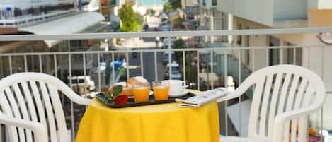 Un balcón con una mesa amarilla lista para el desayuno, sillas blancas y vistas a una calle y edificios de la ciudad.