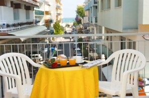 A balcony with a yellow table set for breakfast, white chairs, and a view of a city street and buildings. - Hotel Confort (Cattolica)