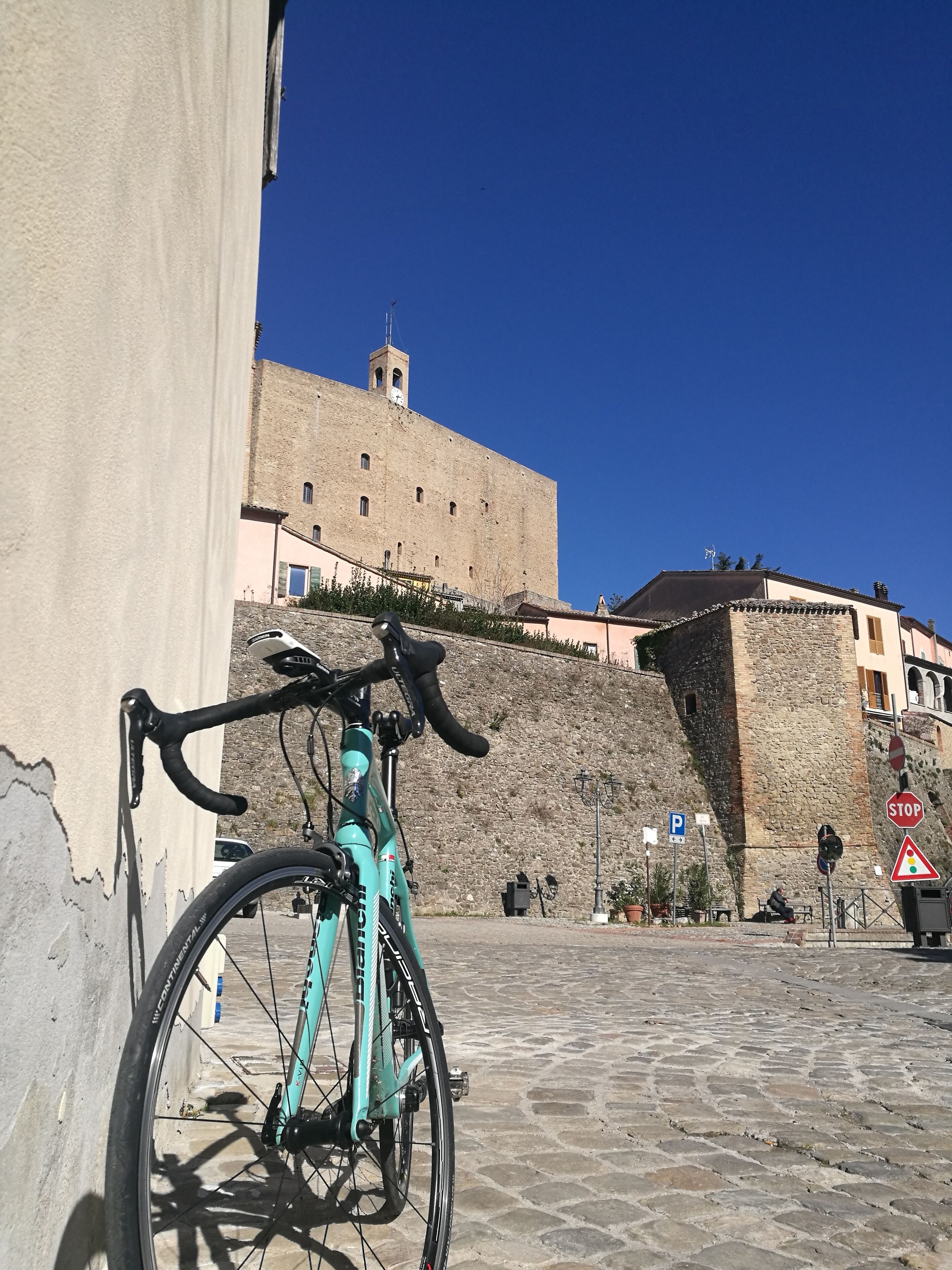A turquoise bicycle parked against a wall, with a historic stone building and a clear blue sky in the background.