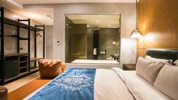 Une chambre d’hôtel moderne dotée d’un grand lit, d’une tête de lit en bois, d’une armoire et d’une salle de bain visible à travers une paroi en verre.
