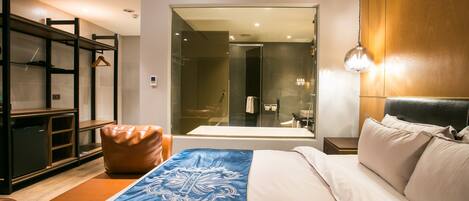 Una habitación de hotel moderna con una cama grande, un cabecero de madera, un armario y un baño visible a través de un mamparo de vidrio.