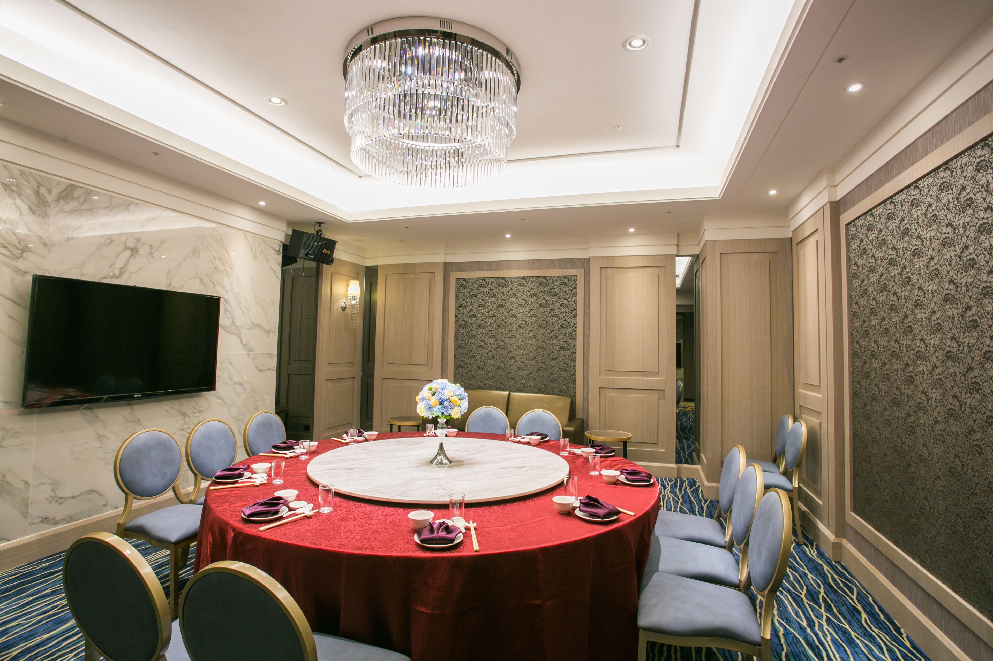 A conference room with a round table, chairs, a chandelier, a TV, and a floral centerpiece.