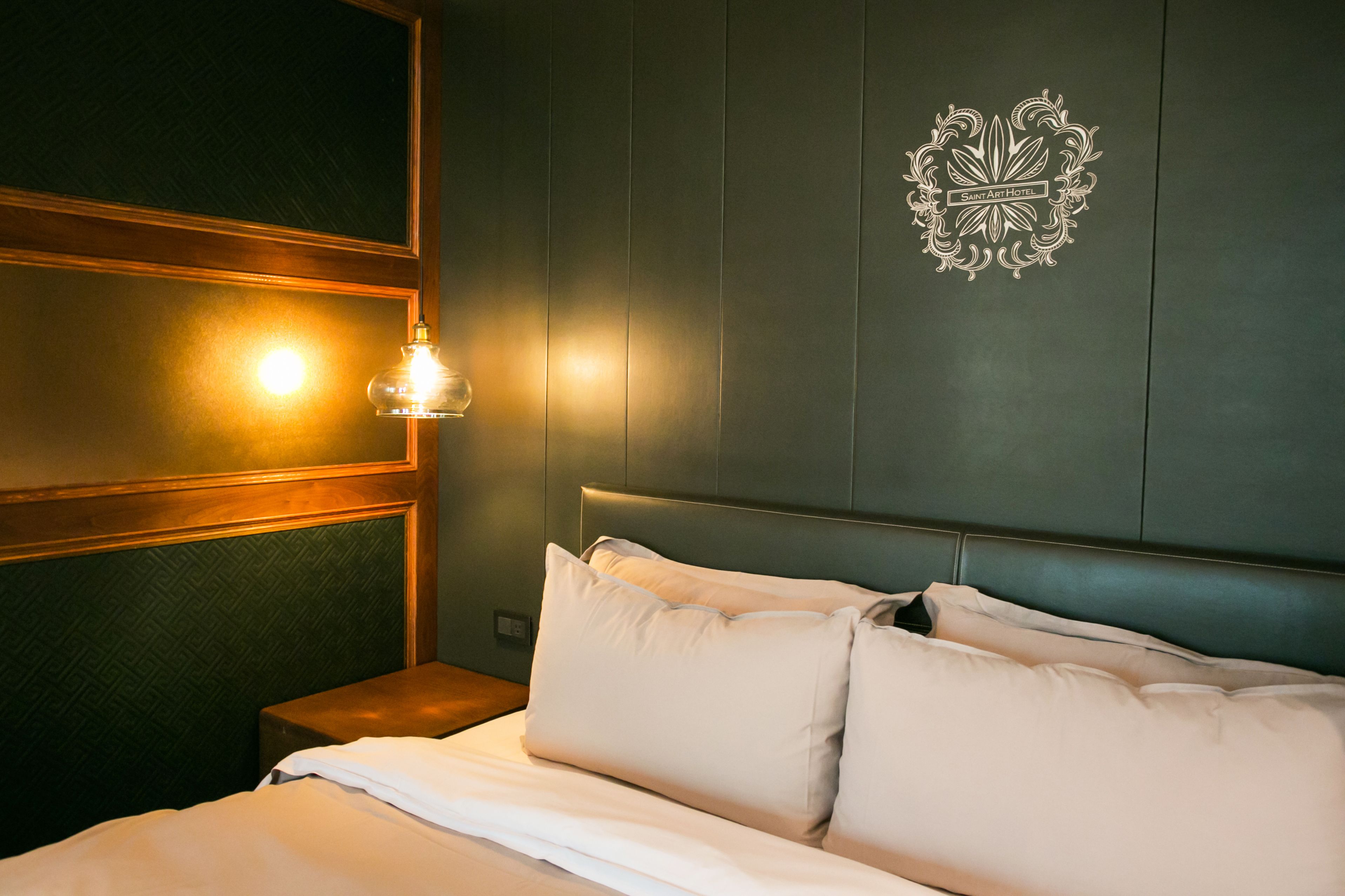 A hotel room with a bed, a nightstand, a wall-mounted light fixture, and a decorative emblem.