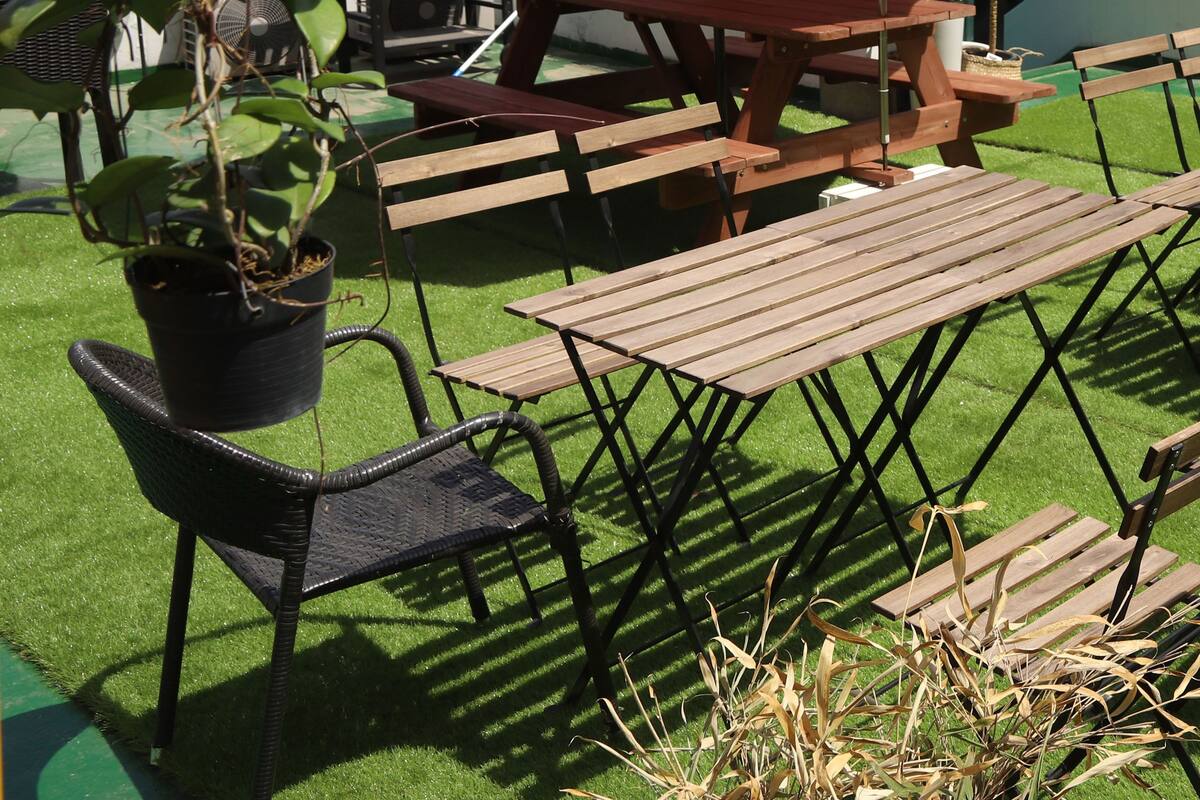 An outdoor patio with wooden tables and benches, surrounded by potted plants and an orange umbrella.