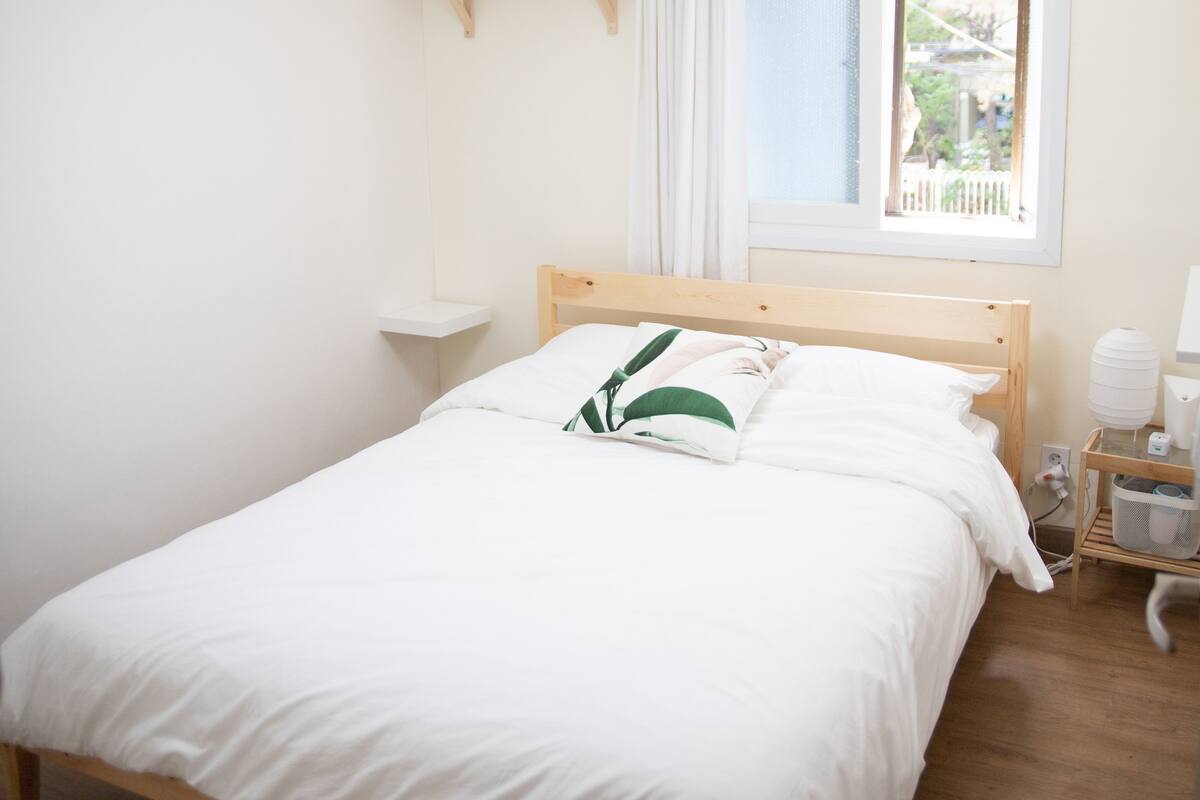 A neatly made bed with white bedding and a green pillow, a bedside table with a lamp, and a window with a view of trees and a fence.