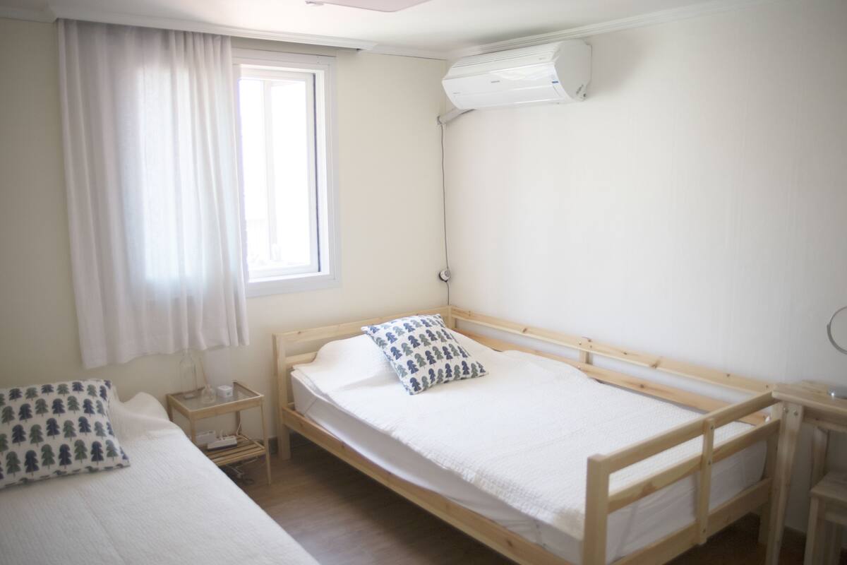 A single-bed room with a wooden frame, white bedding, a bedside table with a lamp, and a window with curtains.
