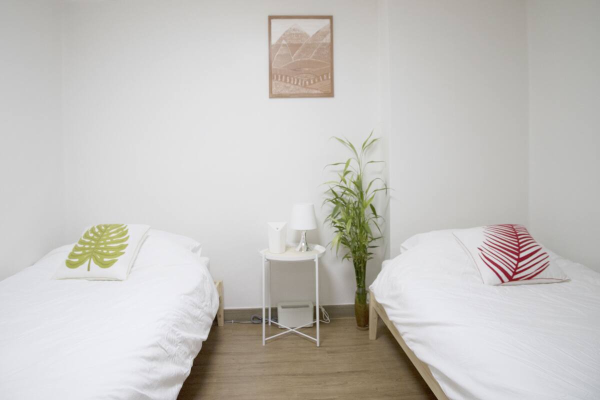 A room with two beds, a small table with a lamp, a potted plant, and a framed picture on the wall.