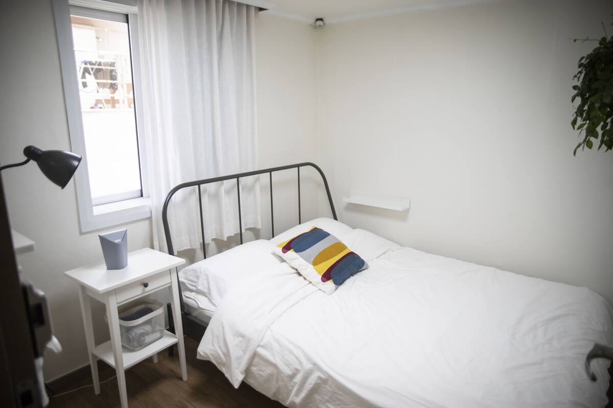 A small, neatly arranged bedroom with a single bed, a bedside table with a lamp, and a window with curtains.