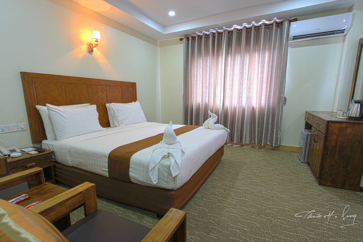 A hotel room with a wooden headboard, a bed with white linens, a bedside table with a phone and remote control, a wooden desk with a chair, a mirror, and a curtain.