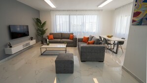 A modern living room with a flat-screen TV, grey sofas with orange cushions, a white coffee table, and a dining area with a white table and chairs.