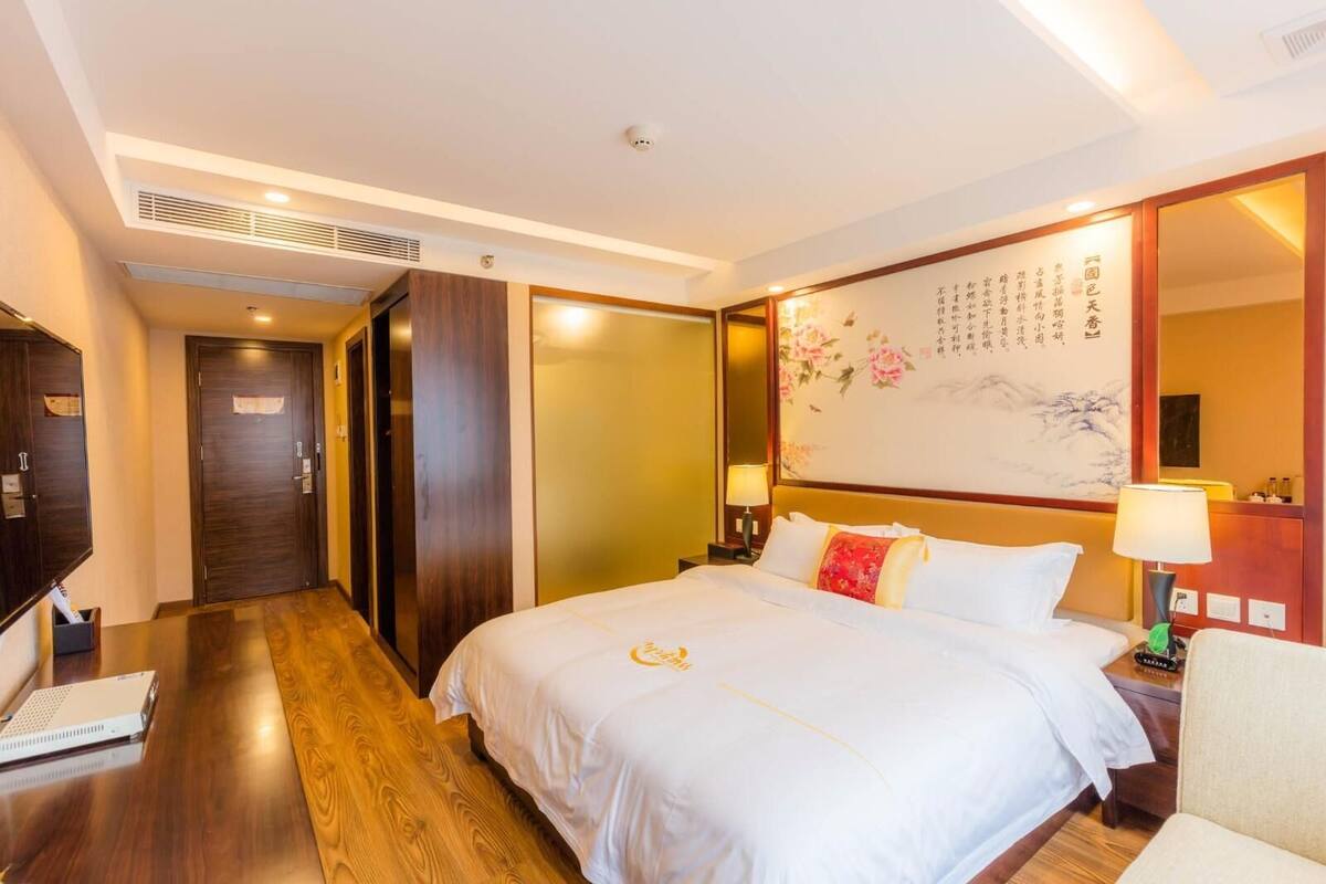 A hotel room with a large bed, wooden flooring, a desk with a laptop, a nightstand with a lamp, and a wall with calligraphy.