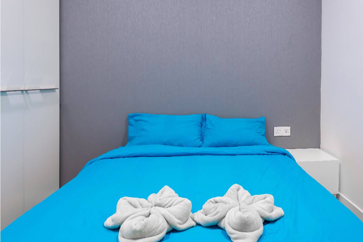 A bed with a blue bedspread and two pillows, a white bedside table, and a grey headboard.