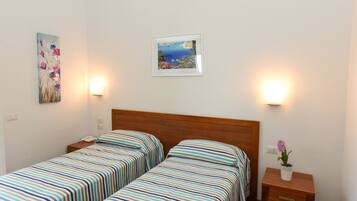 A room with two single beds, a wooden headboard, a nightstand with a phone, a framed picture on the wall, and a small table with a vase of flowers.