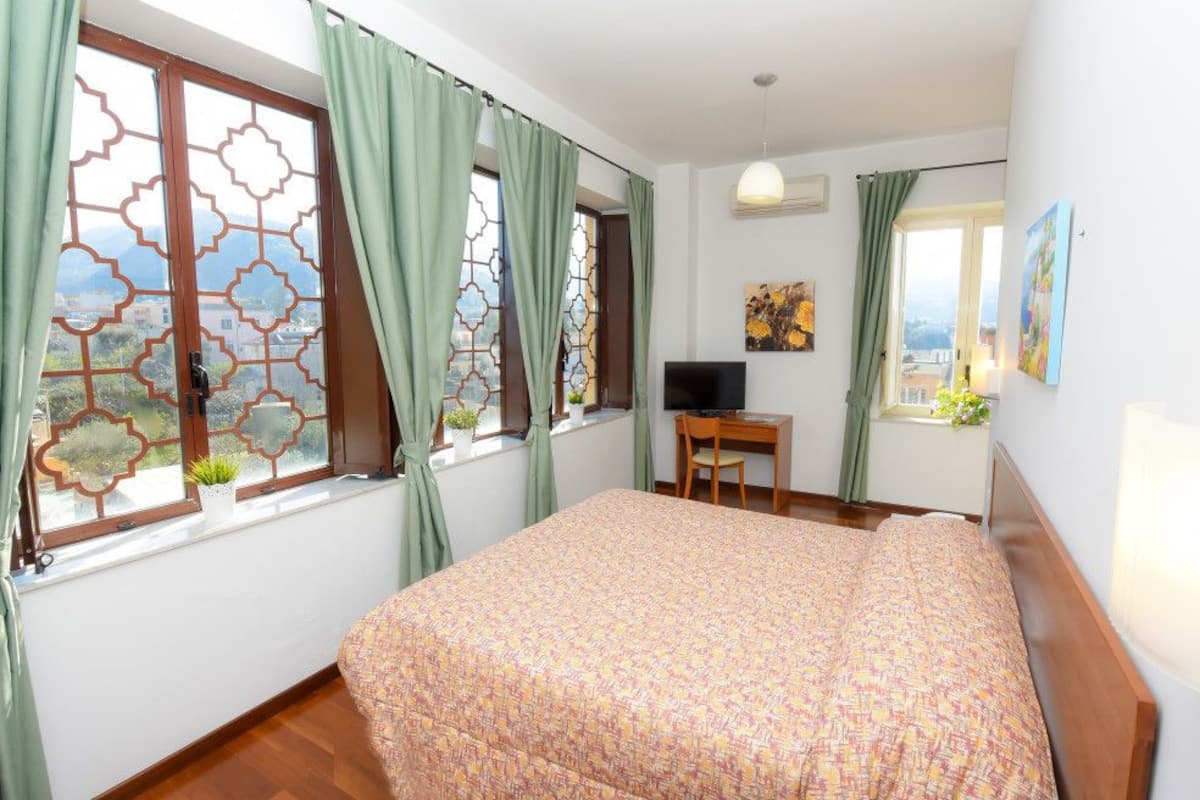A bedroom with a bed, a desk, a TV, and a window with curtains.