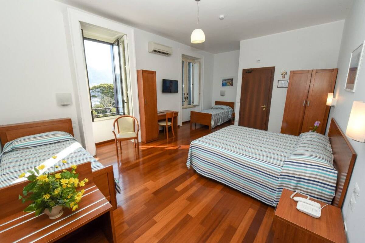 A room with two beds, a desk, and a chair. There is a window with a view of trees.