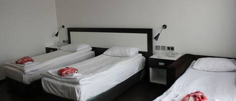 A hotel room with three single beds, each with a pillow and a blanket. There are bedside tables with lamps and a phone.