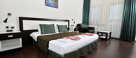 Une chambre d’hôtel bien aménagée, avec un grand lit, une table de chevet, une commode, une chaise, une petite table et une fenêtre avec des rideaux.