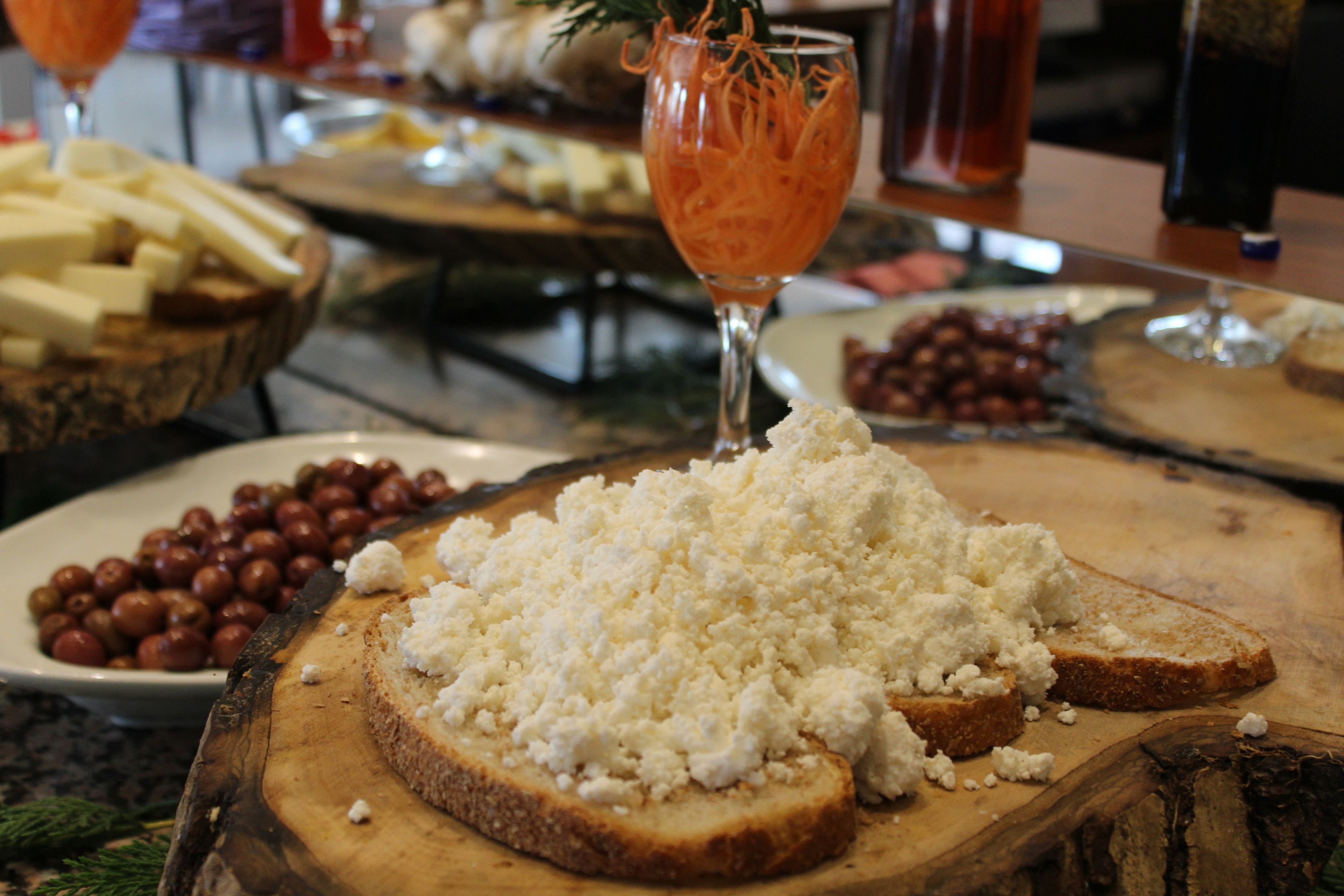 A wooden board with cheese and bread, a glass with a drink, and a bowl of olives.