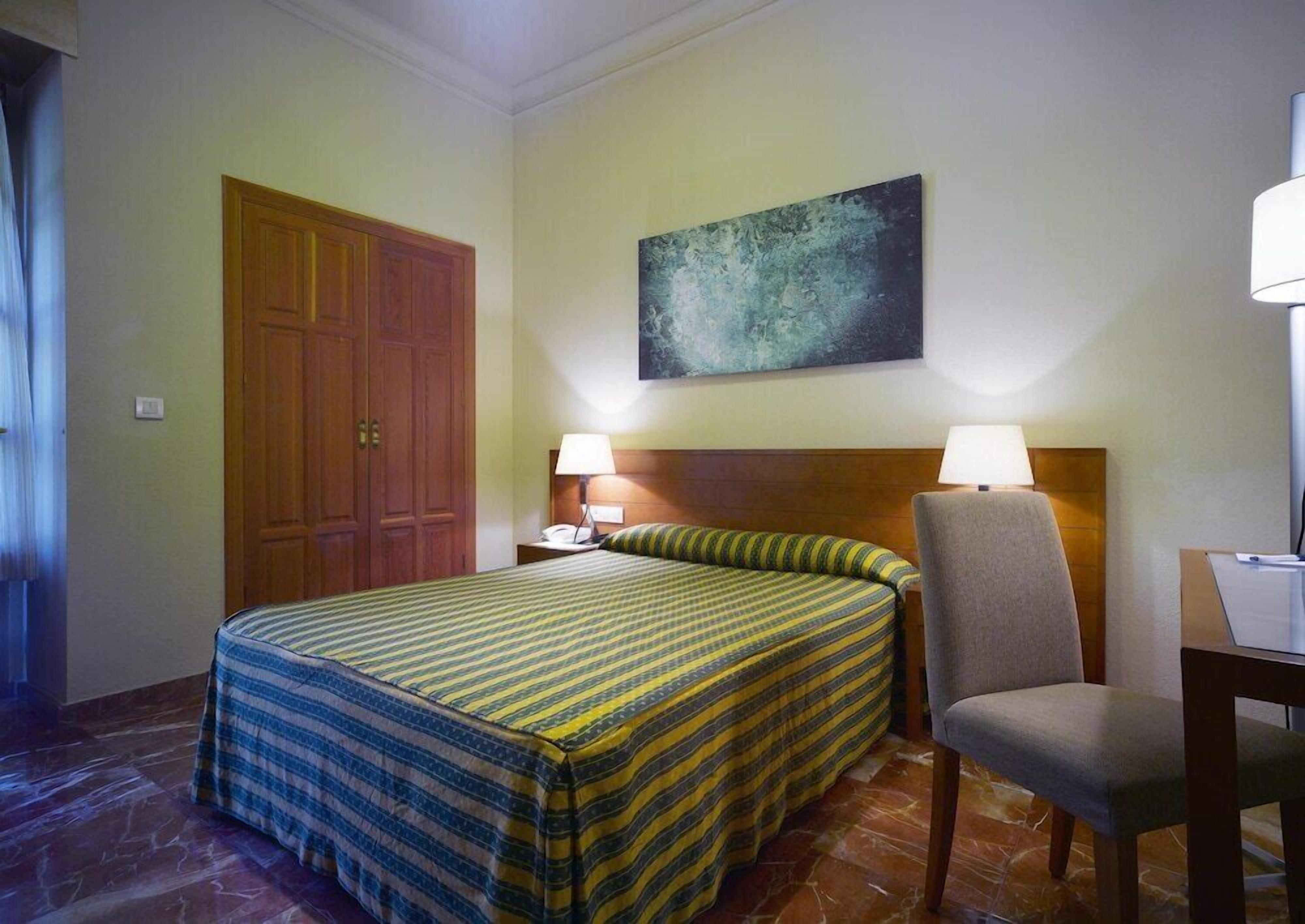 Una habitació d'hotel amb llit, una cadira, escriptori, un quadre a la paret i una porta.