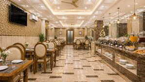 A hotel dining area with multiple tables and chairs, a buffet counter, and a ceiling fan. - OPO Viva Palace (New Delhi)