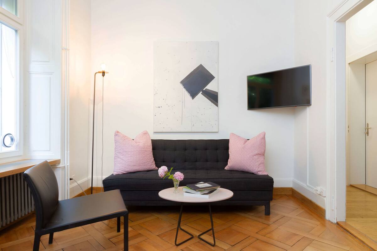 Ein modernes Wohnzimmer mit einem schwarzen Sofa, einem kleinen runden Tisch, einem schwarzen Sessel, einer Stehlampe und einem flachbildfernseher an der Wand.