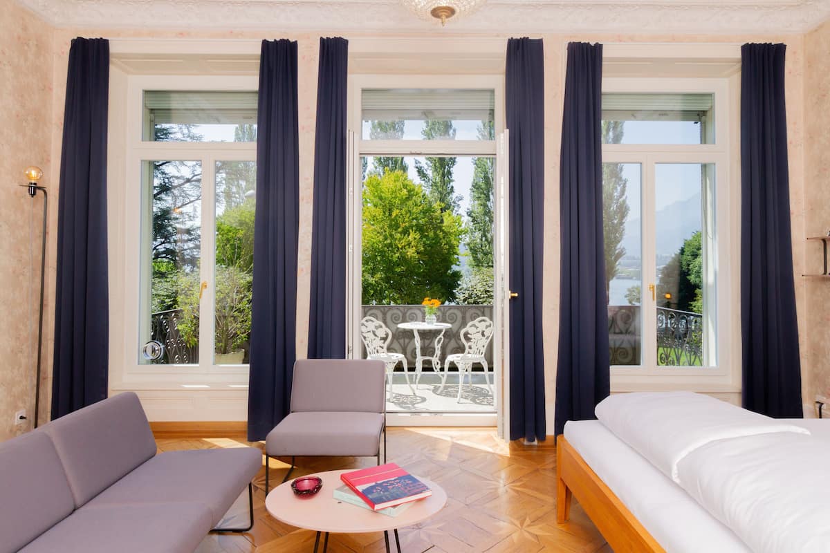 Ein modernes Hotelzimmer mit einer Couch, einem kleinen runden Tisch mit Büchern, einem Bett und einem Balkon mit Gartenmöbeln.