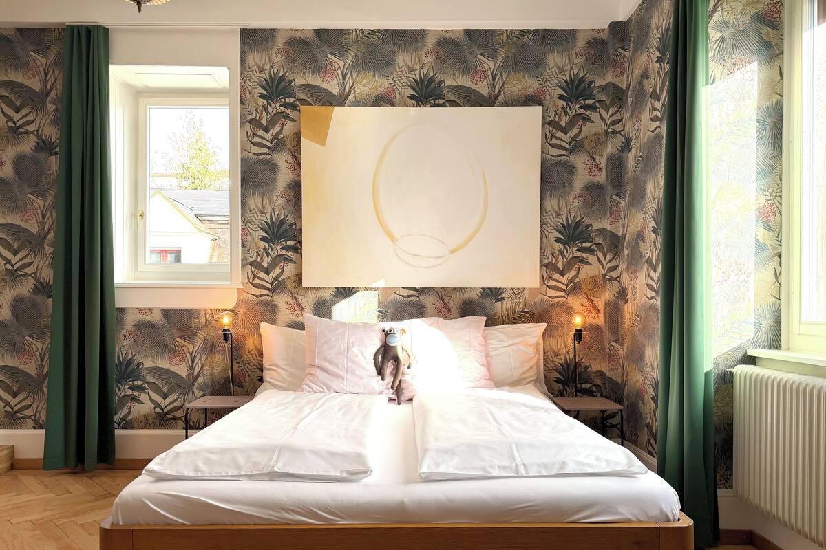 Ein Schlafzimmer mit einem Bett, einem an der Wand hängenden abstrakten Gemälde und einem Fenster mit Vorhängen.