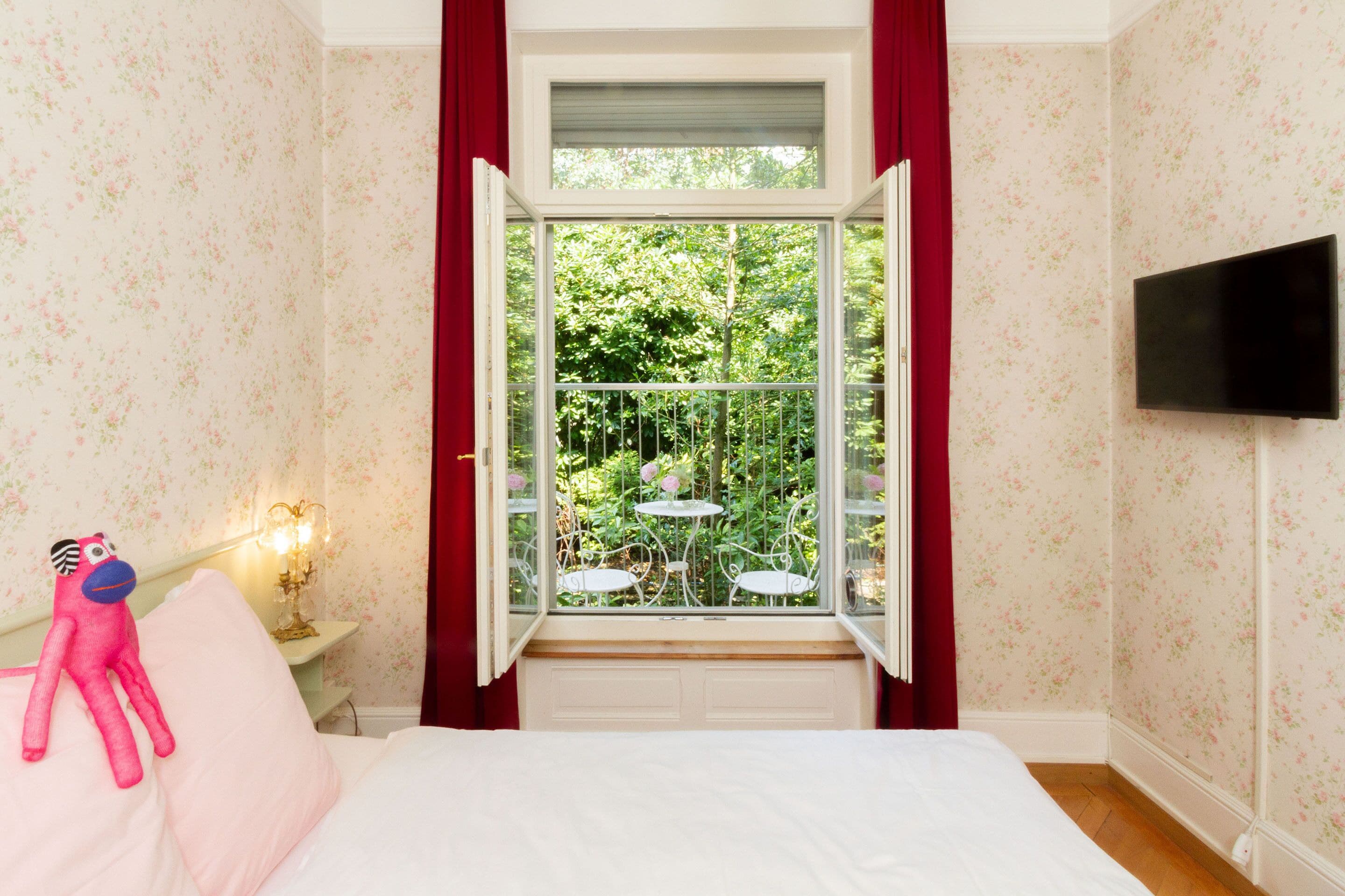 Une chambre à coucher avec un lit, une fenêtre ornée de rideaux rouges, un téléviseur fixé au mur et une petite table avec une lampe.