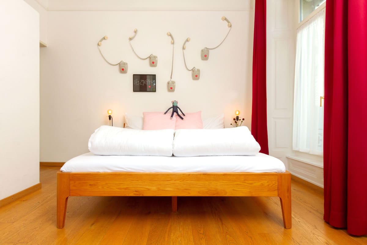 Ein Schlafzimmer mit einem hölzernen Bett, weißer Bettwäsche, einer Wanduhr und einem roten Vorhang.