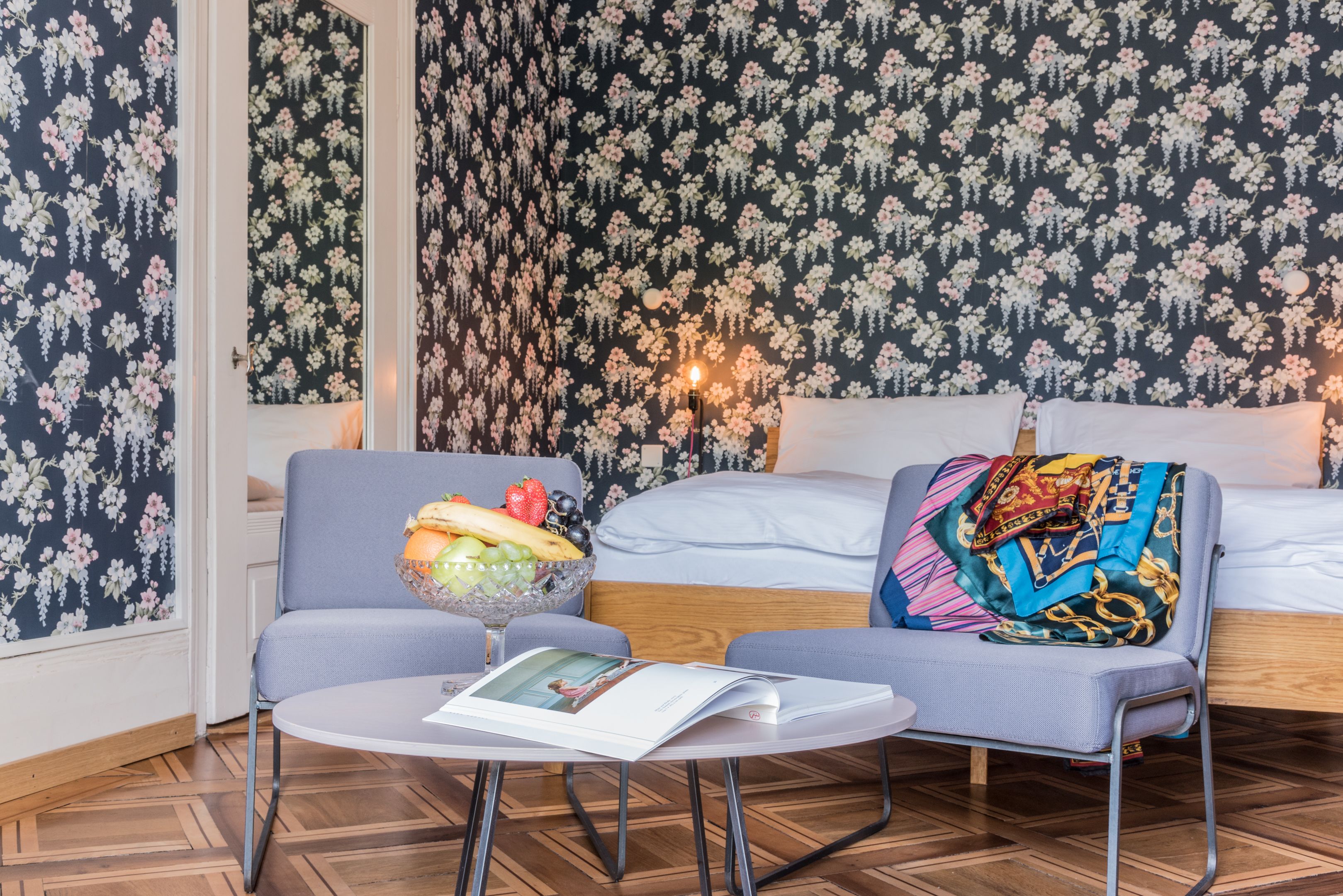 A cozy room with a floral wallpaper, a sofa with a colorful blanket, a small table with a magazine, and a fruit bowl.