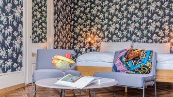 A cozy room with a floral wallpaper, a sofa with a colorful blanket, a small table with a magazine, and a fruit bowl.