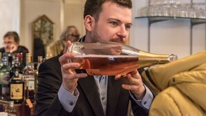 Una persona sostiene una botella grande, probablemente de vino o champán, en un bar con varias botellas y copas de fondo.