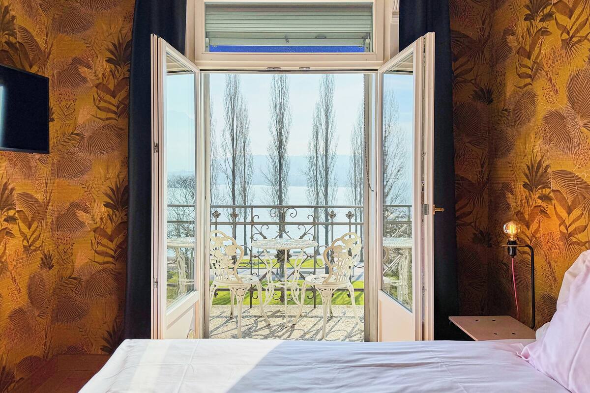 Ein Schlafzimmer mit Bett, Nachttisch, Lampe und einem Balkon mit Stühlen und Tisch.