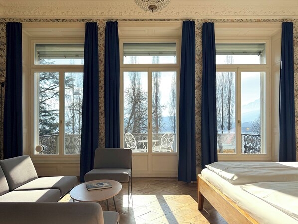Habitación de hotel con cama, dos sillones, una mesita, ventanas grandes con cortinas y un balcón con barandilla.
