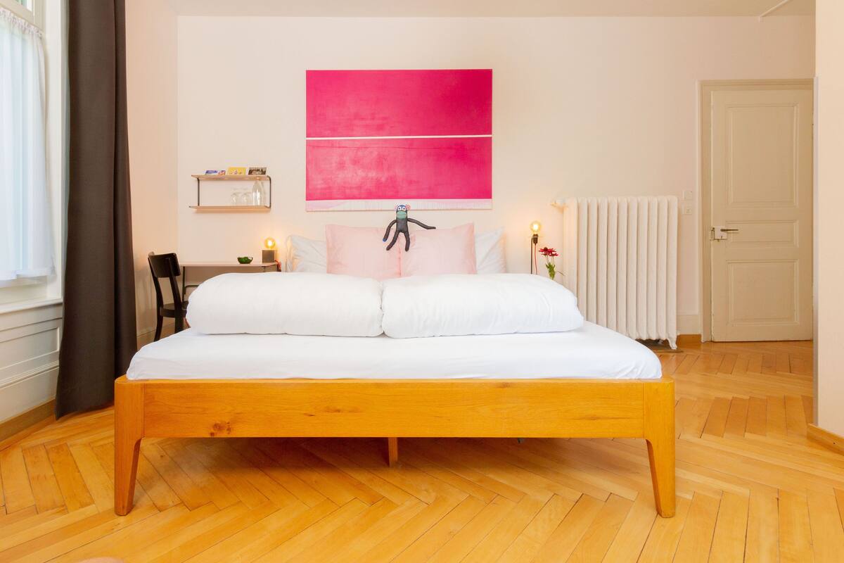 Ein Schlafzimmer mit einem Holzbett, weißer Bettwäsche, einem abstrakten rosa Gemälde, einer Heizung und einer Tür.