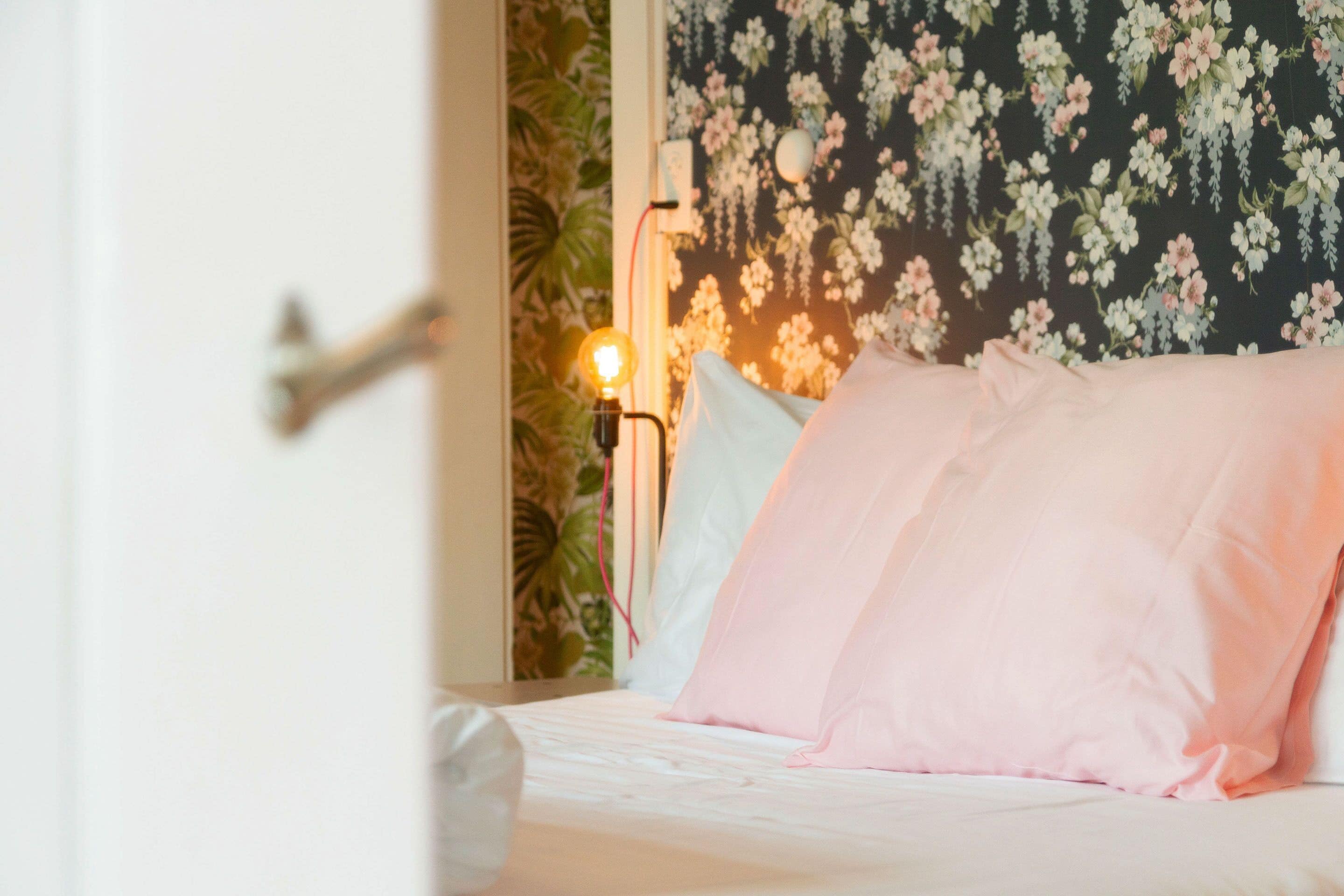 Un dormitorio con papel tapiz floral, una cama con ropa de cama blanca y rosa, y una lámpara de noche.