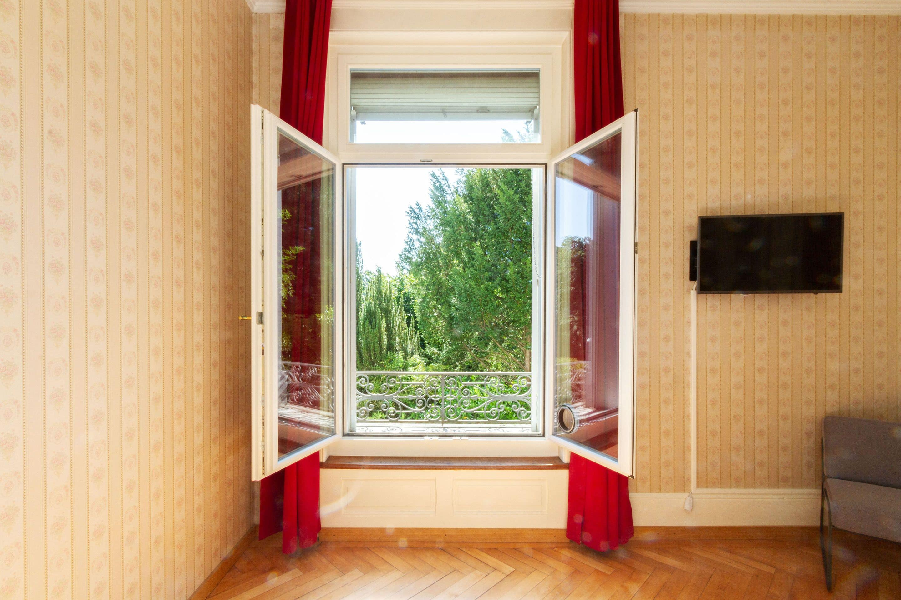 A room with a large window, wooden flooring, and a television on the wall.