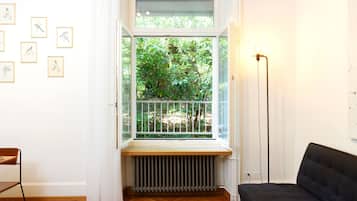 Quarto com uma janela grande, um radiador, um sofá preto, um abajur e quadros nas paredes.