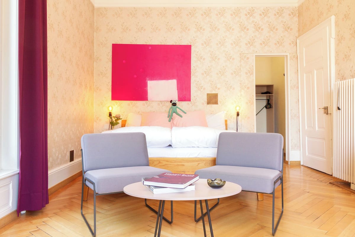 Ein Hotelzimmer mit einem Bett, zwei Sesseln, einem kleinen Tisch mit Büchern, einem roten Kunstwerk an der Wand und einer Tür zu einem weiteren Raum.