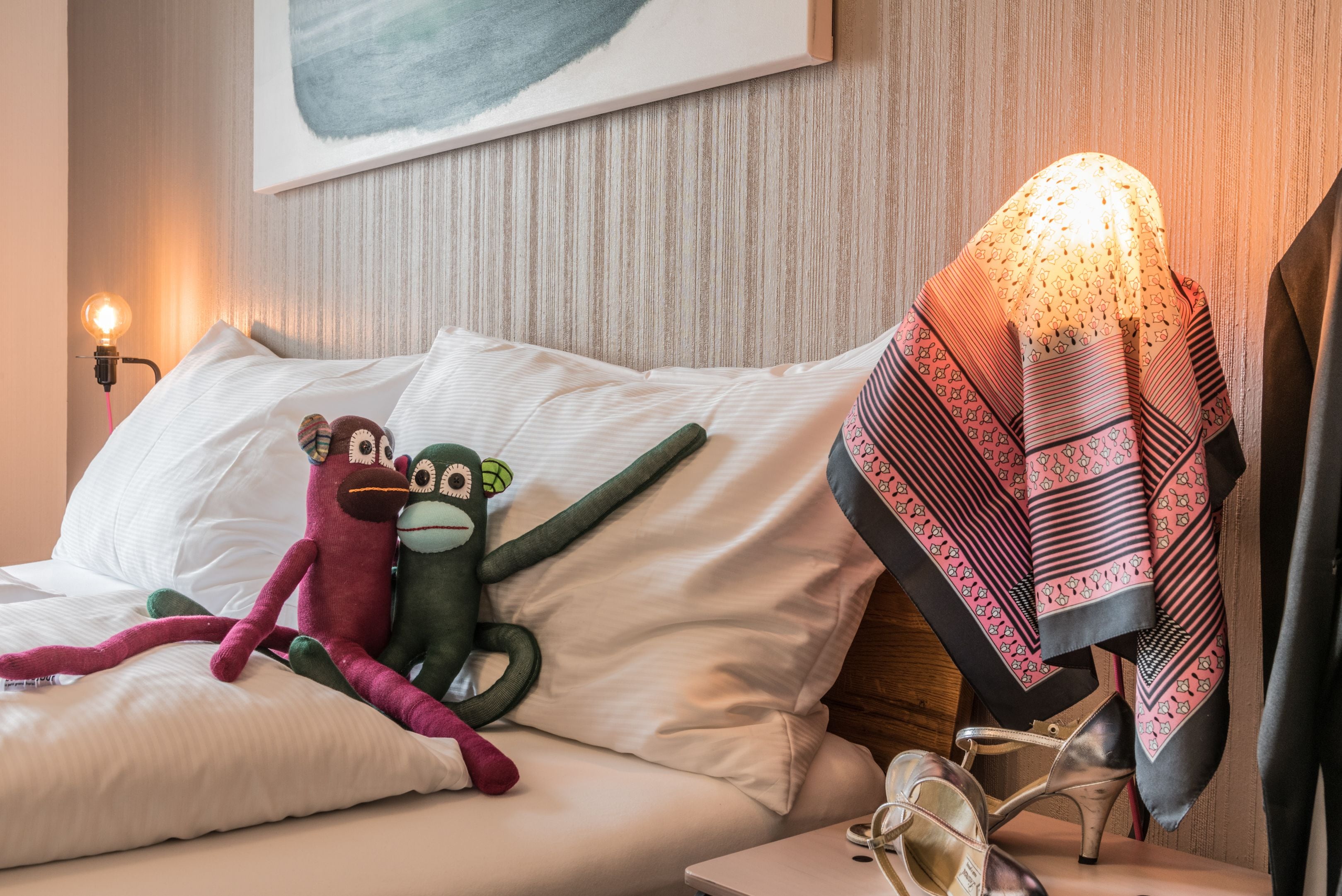 Un lit avec un jouet en peluche, une lampe et une paire d’escarpins sur une table de chevet.