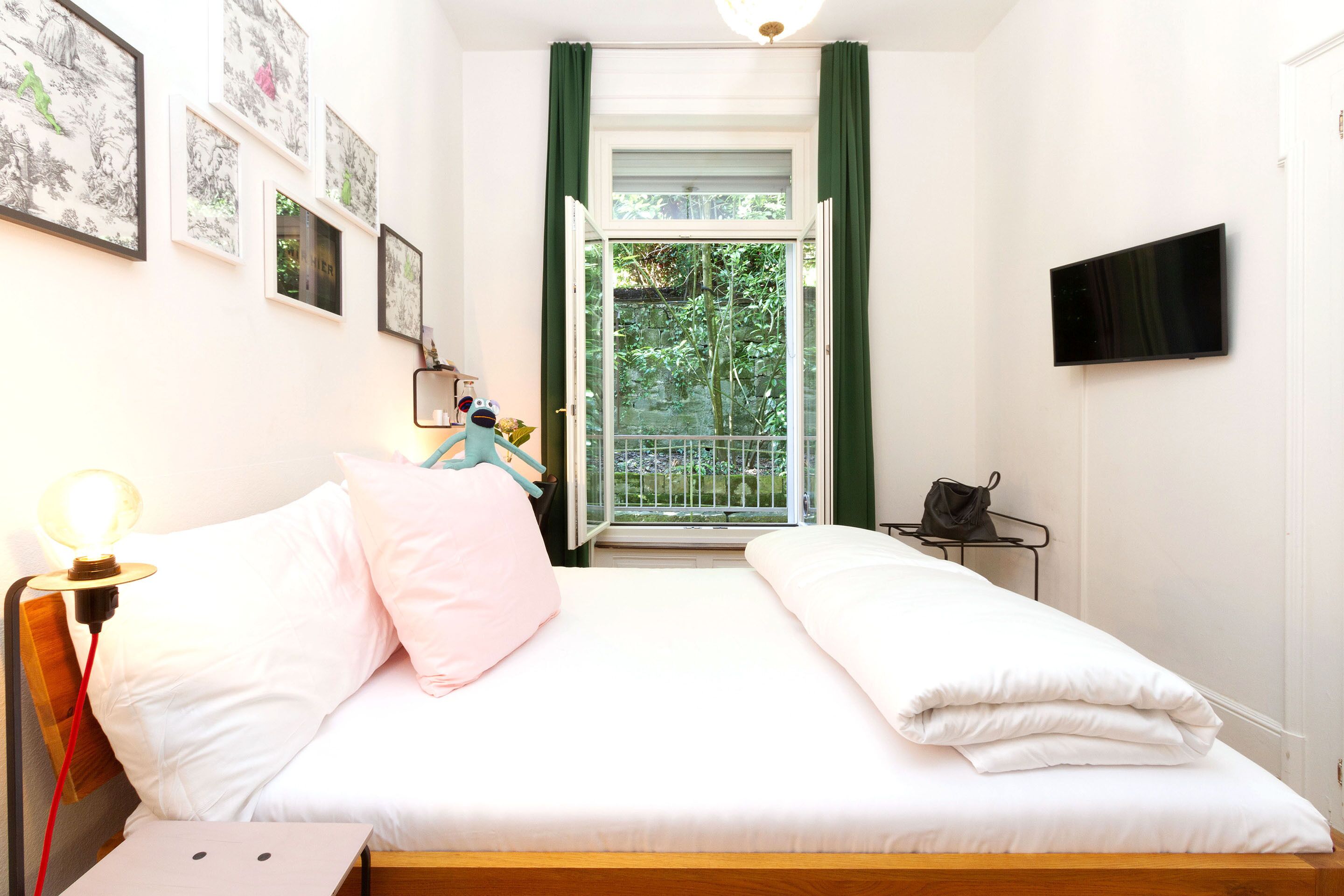 Un dormitorio con una cama, una mesita de noche, una lámpara, un televisor y una ventana con cortinas verdes.