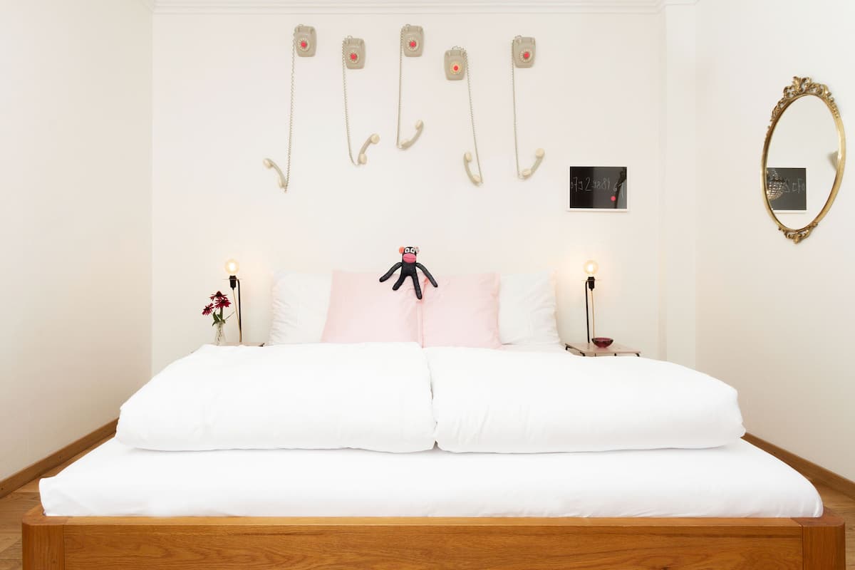 Ein Schlafzimmer mit einem hölzernen Bett, weißer Bettwäsche, einem Stofftier auf den Kissen, einem an der Wand befestigten Telefon, einem Spiegel und einem kleinen Tisch mit einer Lampe.