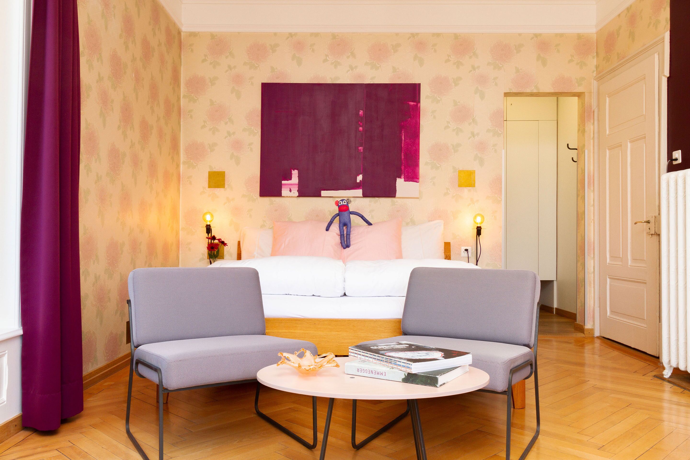 Ein Hotelzimmer mit einem Bett, zwei Sesseln, einem runden Tisch mit Büchern und einem großen abstrakten Gemälde an der Wand.