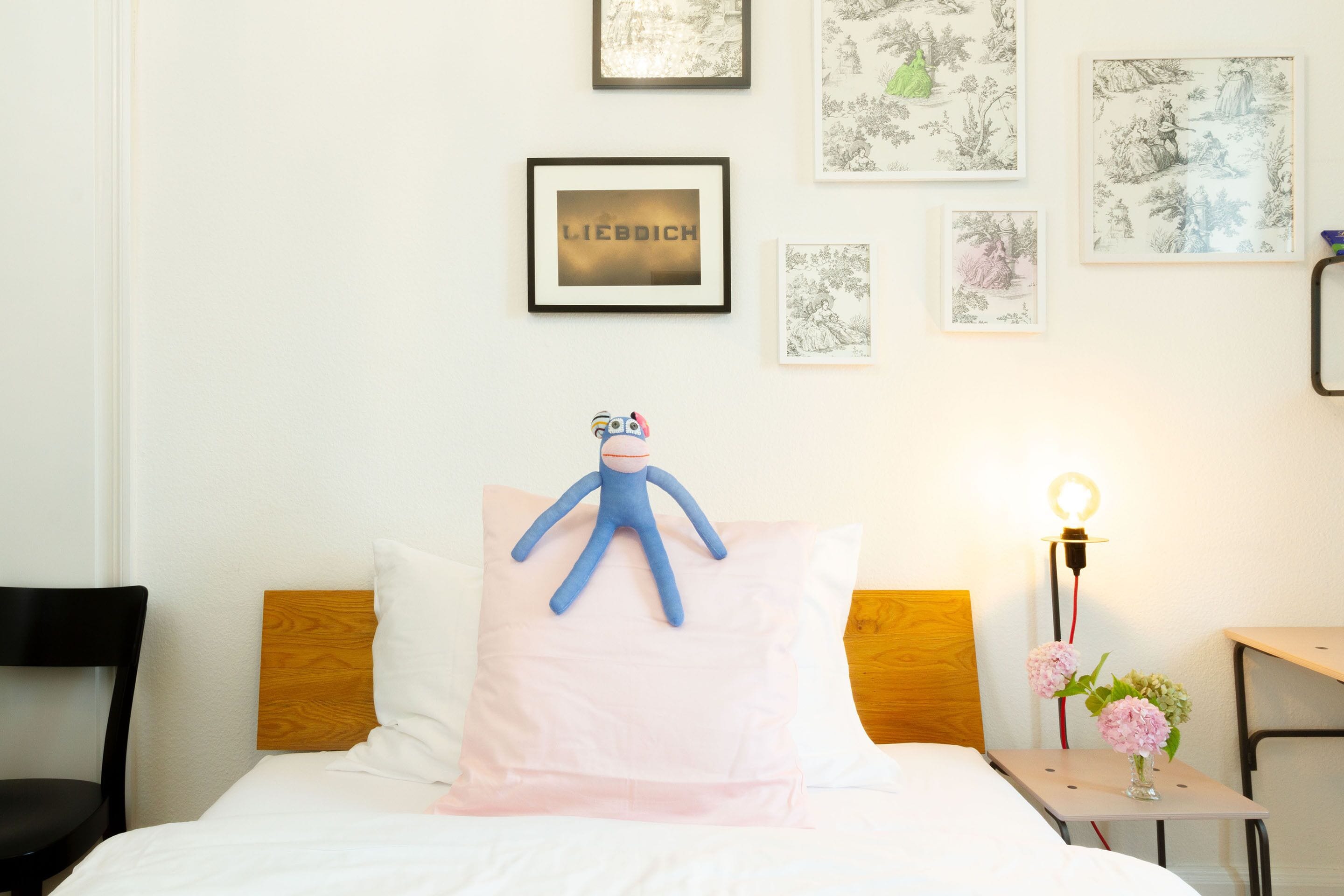 A neatly made bed with a blue plush toy on a pink pillow, a small bedside table with a lamp and flowers, and framed artwork on the wall.