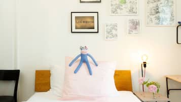 Una cama bien hecha con un peluche azul sobre un almohadón rosa, una mesita de noche con una lámpara y flores, y cuadros en la pared.