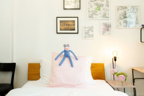 A neatly made bed with a blue plush toy on a pink pillow, a small bedside table with a lamp and flowers, and framed artwork on the wall.