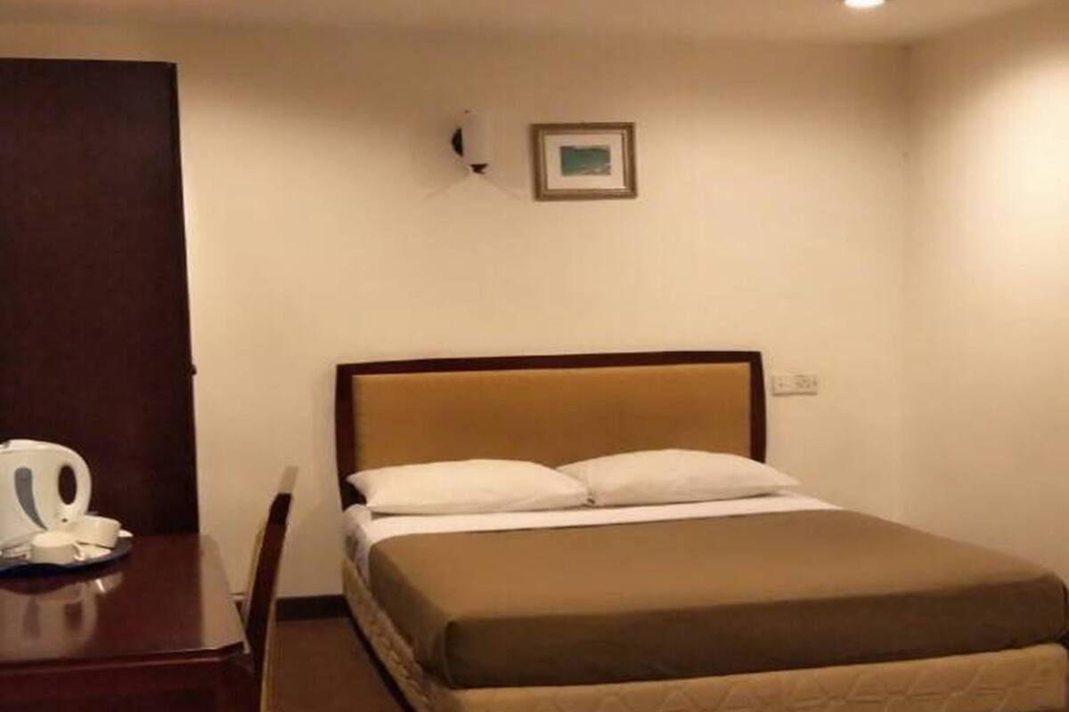 A hotel room with a single bed, a wooden nightstand, a chair, and a desk with a kettle.