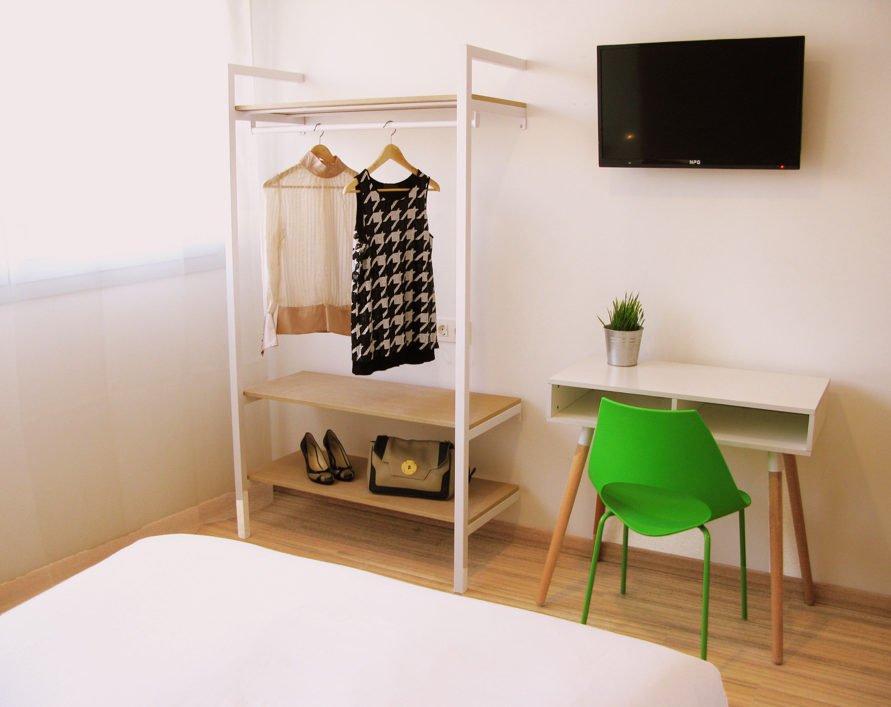 A bedroom with a bed, a green chair, a desk with a plant, a TV, and a wardrobe with clothes and shoes.